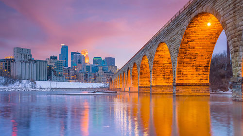 Bridge over river by buildings against sky during sunset