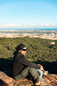Young man sitting on rock against sky