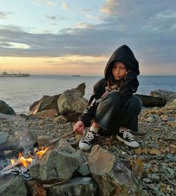 Girl in black hood sitting at beach by the fire during sunset