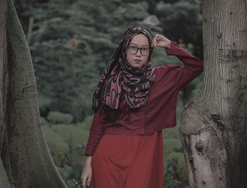 Portrait of young woman wearing hijab standing by tree trunk in forest