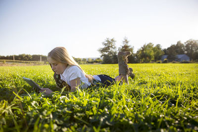 Schoolgirl using tablet computer while lying on grassy field against clear sky