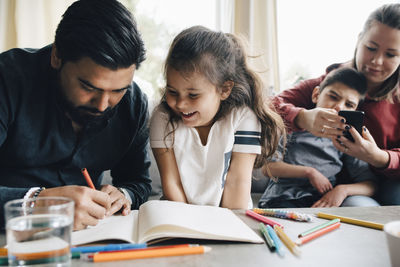 Father teaching drawing to daughter while woman using smart phone with autistic son in background