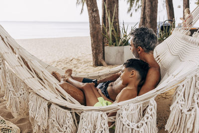 Shirtless father and son relaxing in hammock at beach on vacation