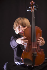 Boy holding violin while sitting against black background