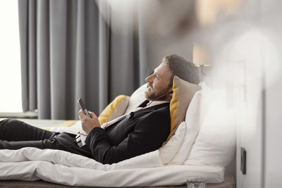 Side view of businessman listening to music while lying on bed at hotel room