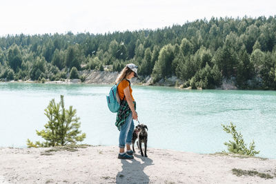 Side view of young woman traveler with backpack walking on sand shore of blue lake or river with dog