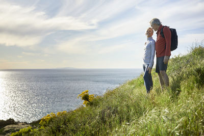 Senior couple talking while standing on cliff by sea against sky