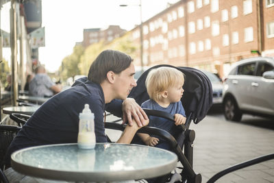 Father and son looking away while sitting at sidewalk cafe in city