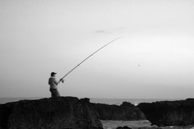 Man fishing on rock by sea against sky