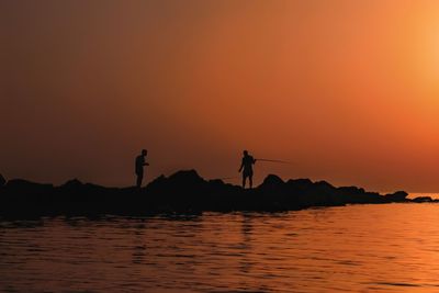 Silhouette people standing on shore against orange sky