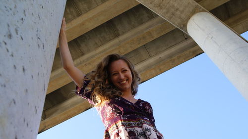 Low angle portrait of smiling young woman standing below bridge