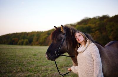 Beautiful young woman standing by horse on field during sunset