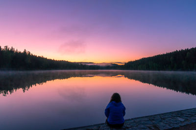 Rear view of woman sitting at lake against sky during sunset