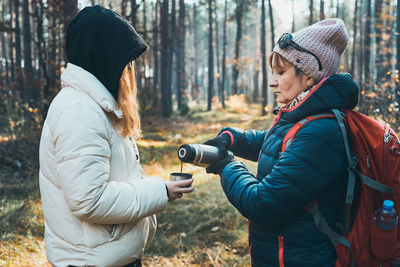 Mother and daughter with backpack having break during autumn trip pouring a hot drink from thermos