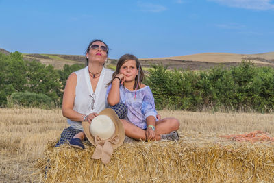 Portrait of mother and daughter sitting on hay bale against blue sky