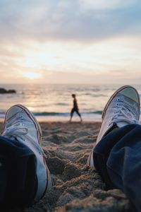 Low section of man sitting on sand while woman walking in background at beach during sunset