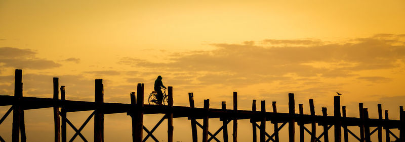 Silhouette man riding bicycle on pier against orange sky during sunset