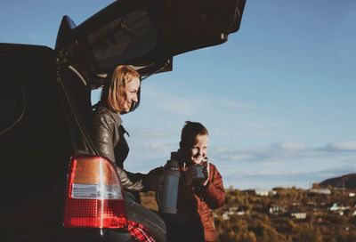 Happy young woman standing by car against sky