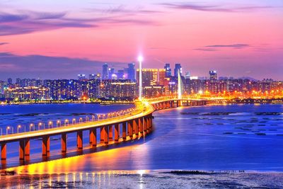 Illuminated bridge over river by buildings against sky at night