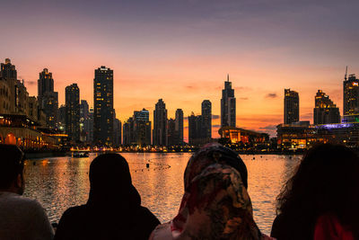 Rear view of people by river in city during sunset