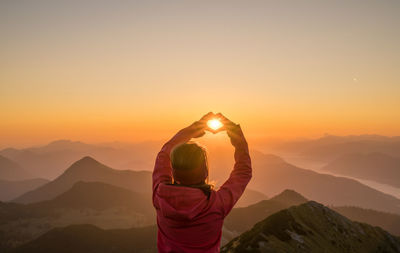 Rear view of girl gesturing heart shape against mountains during sunset