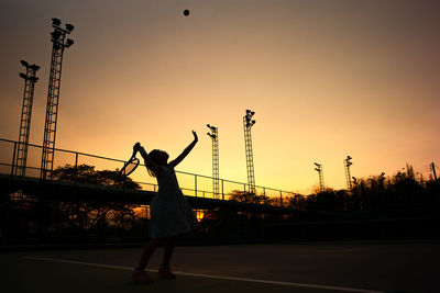 Silhouette girl playing tennis while standing in court against sky during sunset