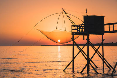 Silhouette fishing net by sea against orange sky