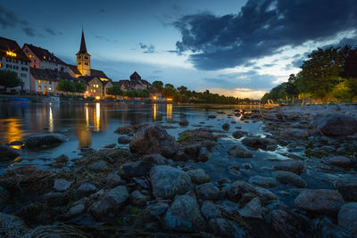 River by illuminated buildings against sky at dusk