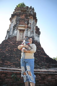 Portrait of woman carrying dog while standing against old ruin