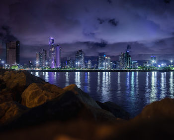 Illuminated city by sea against sky at night