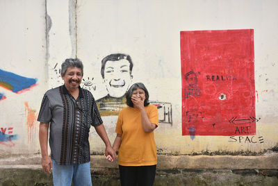 Portrait of mature couple holding hands while standing against graffiti wall