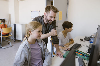 Smiling male teacher assisting female student using computer in classroom at school