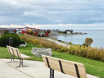 Empty chairs in park overlooking community by lake against sky