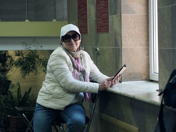 An elderly woman in a white jacket sits in the sanatorium hall   and uses a cell phone.