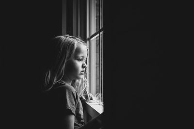 Side view of thoughtful girl looking through window while standing in darkroom at home