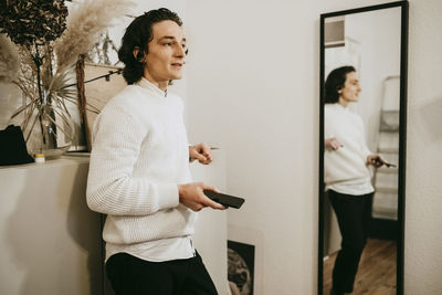 Mid adult man with smart phone standing by mirror at home