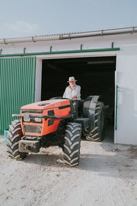 Male farmer in hat driving out of garage in industrial tractor while working in rural area on sunny summer day
