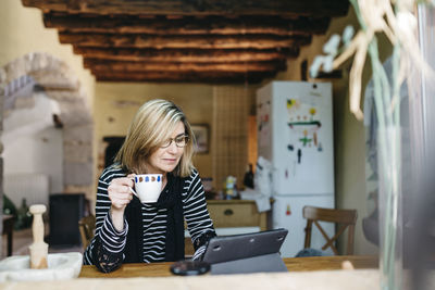 Woman drinking coffee in the kitchen of her farmhouse while using digital tablet