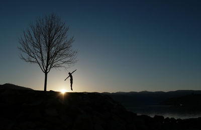 Silhouette boy jumping by bare tree against sky during sunset