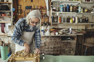 Senior craftsperson working over carved wooden frame at table in workshop