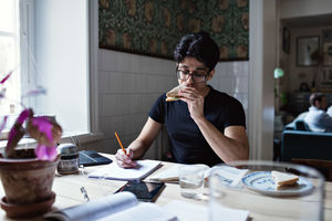 Young man eating sandwich while doing homework at table