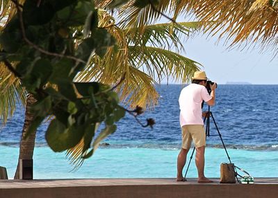 Full length rear view of photographer photographing sea by palm trees