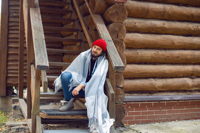 Man with a hipster beard in a red knitted hat and a denim jacket, is stand on stairs at wooden house 