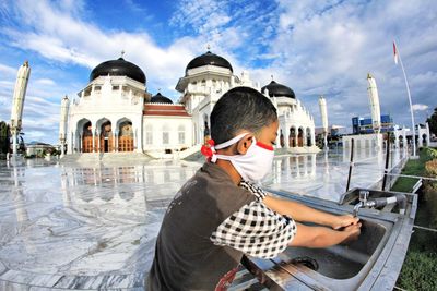 Boy wearing mask cleaning hands at sink against mosque