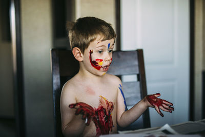 Shirtless messy boy making facial expression while sitting on chair at home