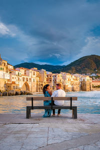 Rear view of people sitting on bench by buildings against sky