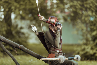 Archer holding bow and arrow while sitting on tree trunk outdoors