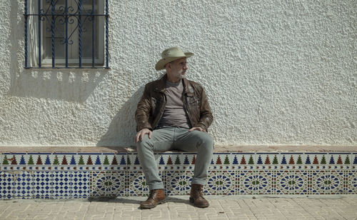 Adult man in cowboy hat sitting against white wall with window