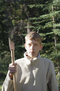 Portrait of teenage boy holding axe against trees at forest