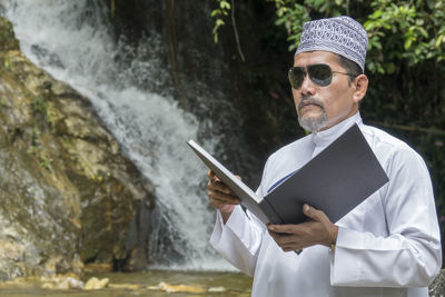 Man reading book while standing against waterfall